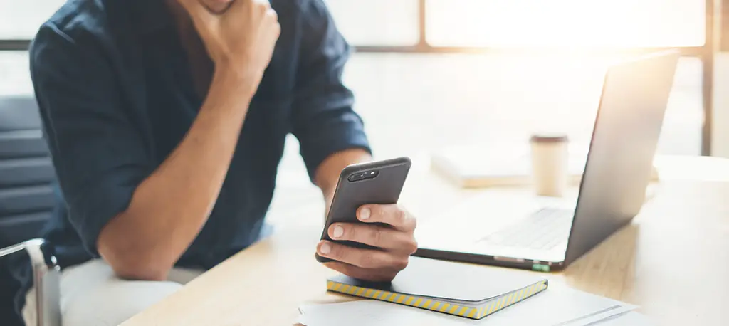 Man looking at his smartphone while sitting in front of a laptop.