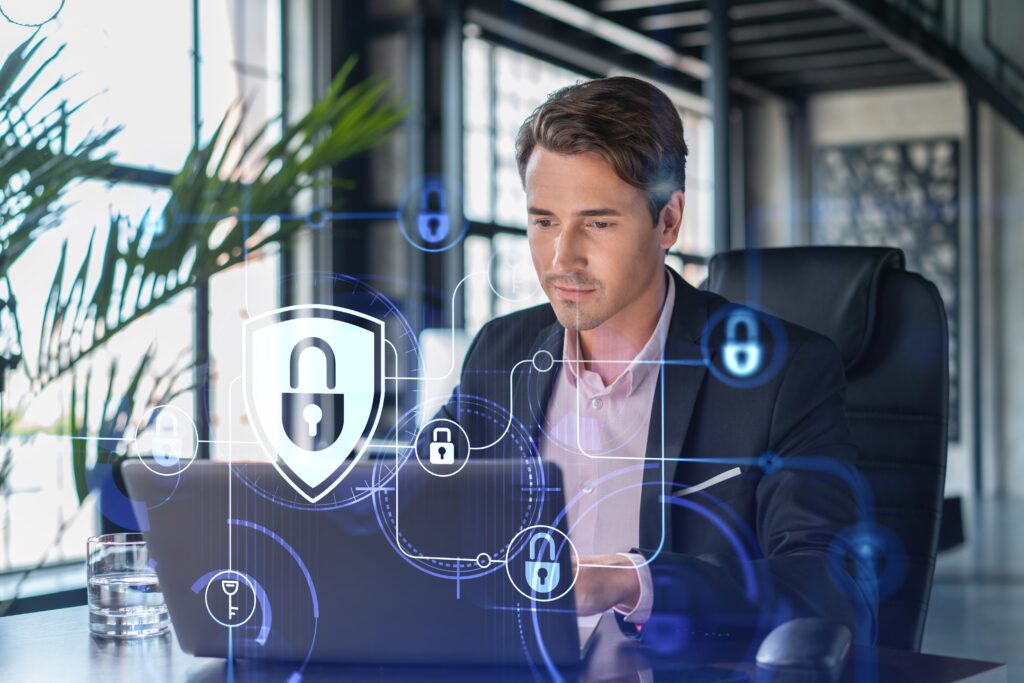 A concept of cybersecurity shown as a padlock hologram over a man working on his laptop in an office