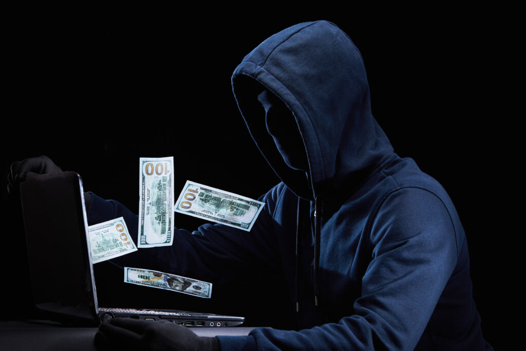 Hooded figure in dark clothing using a laptop, with floating dollar bills suggesting stolen data being turned into profit through cybercrime or data resale